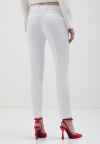 Donna che indossa pantaloni aderenti bianchi e scarpe con tacco rosso a ciabatta, in piedi su uno sfondo bianco uniforme.