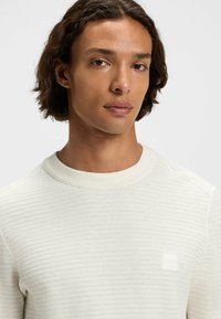 Suéter texturizado blanco con cuello redondo, que presenta un pequeño parche de logo blanco en el pecho y puños acanalados.