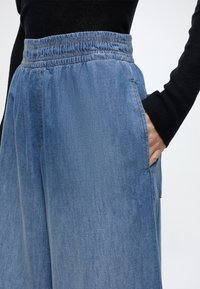 Personne portant un pantalon en denim bleu ample avec une taille élastique, main placée dans la poche latérale, et un haut noir à manches longues visible.