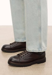 Pantofi din piele maro cu accente texturate, design cu sireturi și talpă groasă din cauciuc negru, asortați cu pantaloni din denim de culoare albastru deschis.