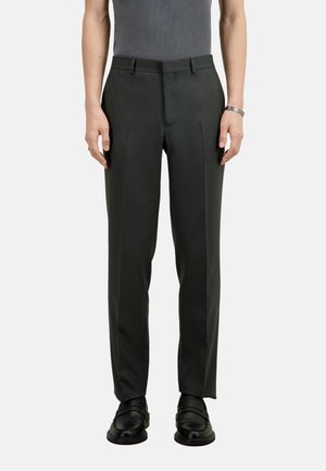 Dunkelgrüne maßgeschneiderte Hose mit geradem Schnitt, einzelnen Falten und Gürtelschlaufen. Getragen mit schwarzen Slippers und einem grauen Oberteil.