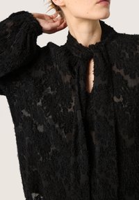 Donna che indossa una blusa sheer nera a trama con scollatura a oblò, sollevando un braccio per toccarsi i capelli su uno sfondo neutro.