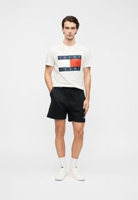 Λευκό βαμβακερό T-shirt με γραφικά σε ναυτικό μπλε και κόκκινο, μαύρα σορτς με κορδόνι και λευκά αθλητικά παπούτσια με λογότυπο. Απλός, καθημερινός σχεδιασμός.