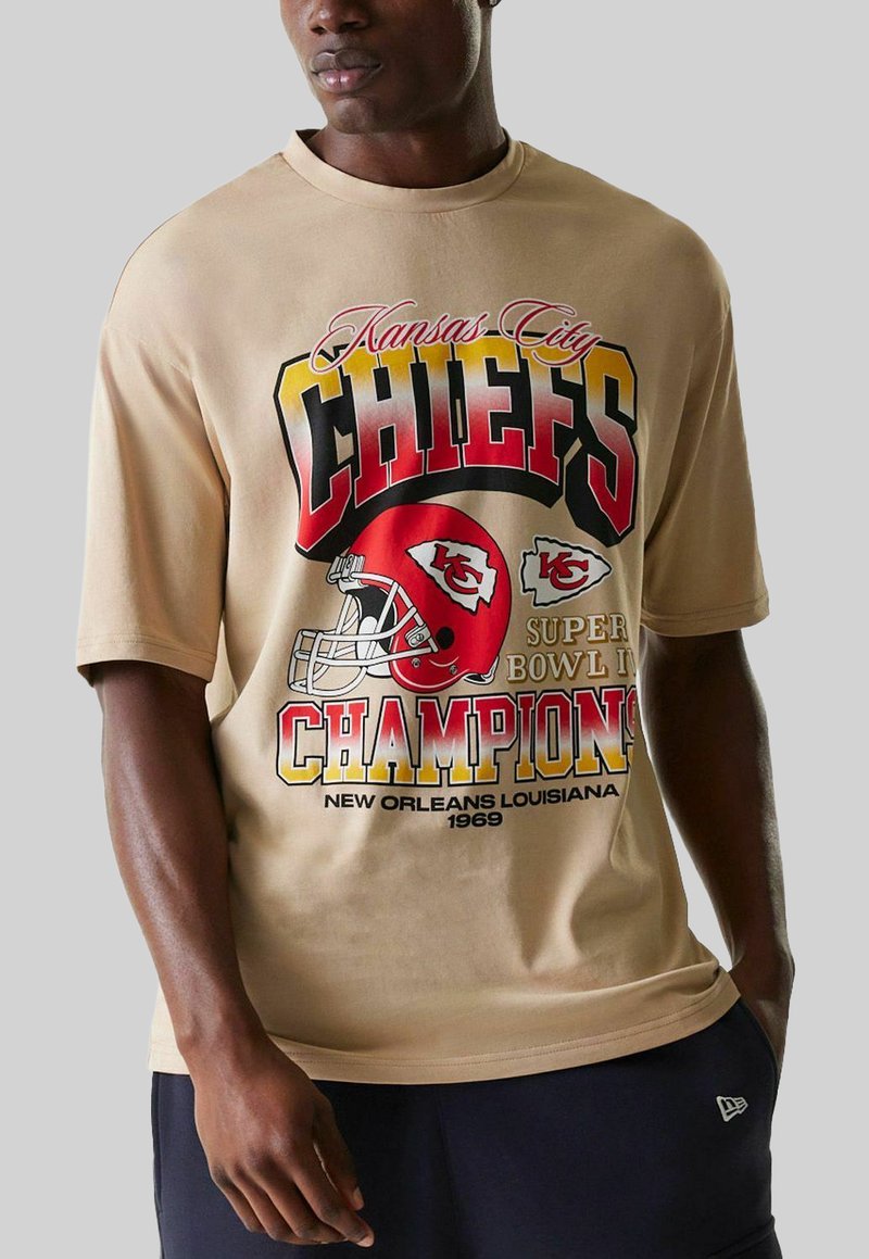 T-shirt beige avec un imprimé graphique d'un casque de football rouge et un texte en gras indiquant "Kansas City CHIEFS CHAMPIONS". Manches courtes, coupe décontractée.
