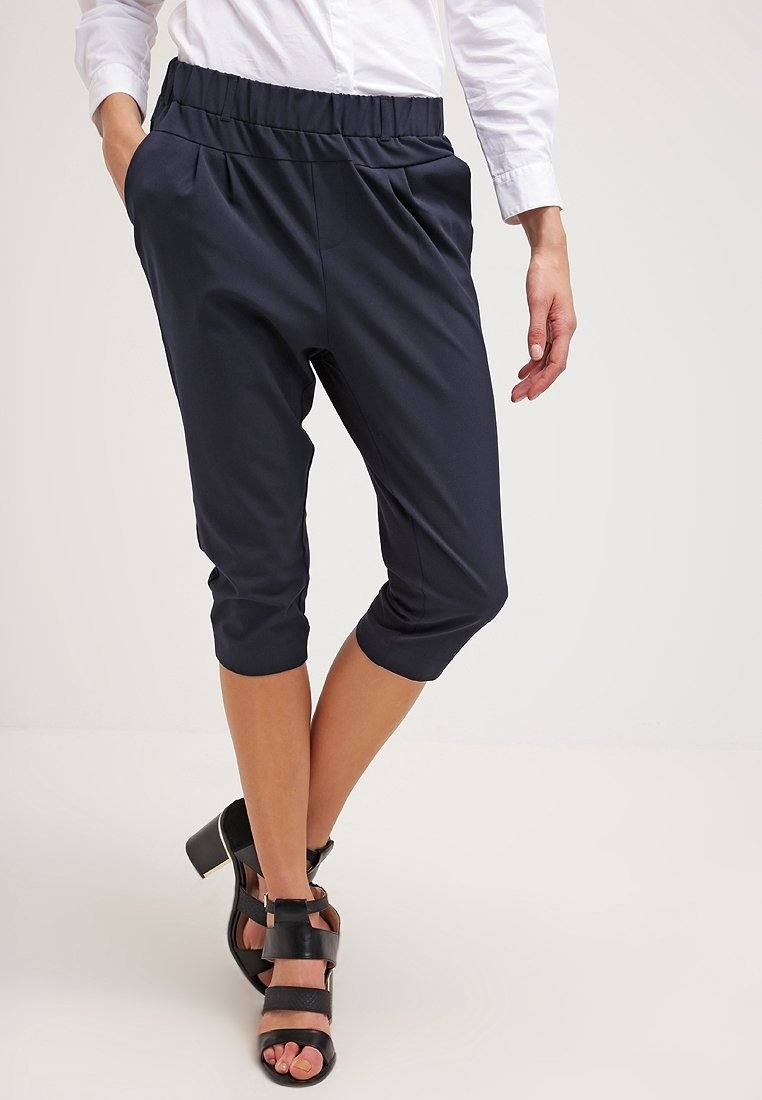 Femme portant un pantalon court bleu marine avec taille élastique, une chemise blanche à manches longues et des sandales noires à talons, debout avec une main dans la poche.