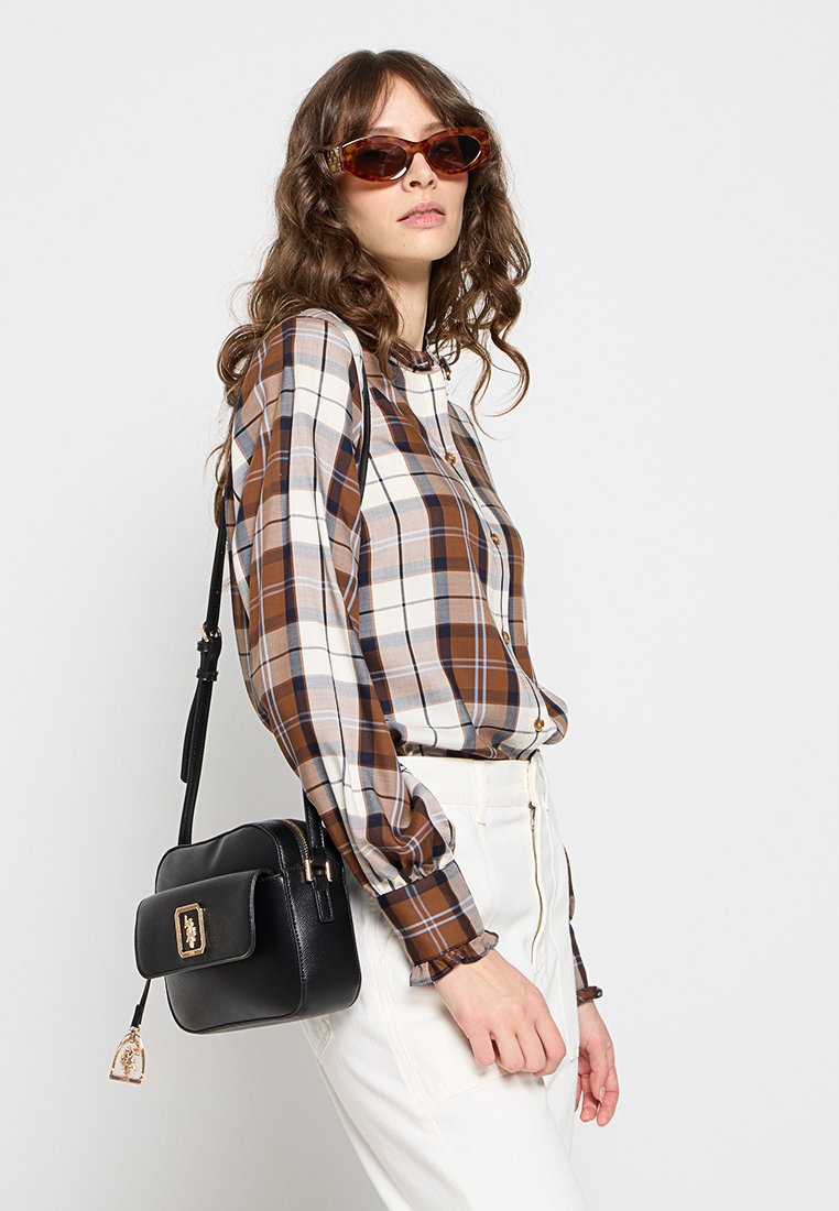 Chemise à carreaux marron et crème avec un devant boutonné, des manches longues et des poignets froncés. Sac bandoulière noir avec quincaillerie dorée. Pantalons blancs.