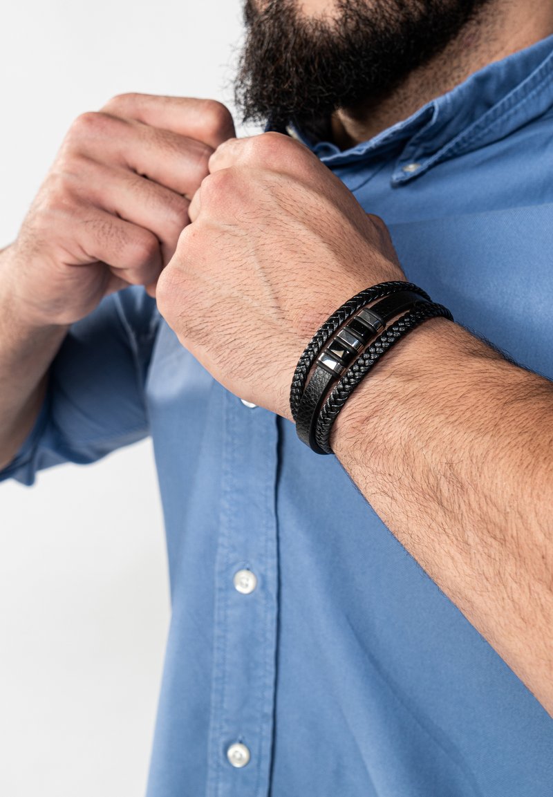 Schwarze geflochtene Armbänder mit glatten, quadratischen Akzenten, die am Handgelenk getragen werden. Die Person passt gerade ein hellblaues, geknöpftes Hemd an.