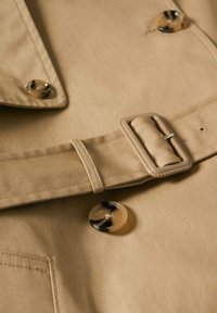 Trench-coat beige boutonné avec une ceinture en tissu et une boucle rectangulaire, gros plan montrant des boutons style écaille de tortue et des détails du col.