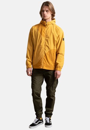 Geel lichtgewicht jack met een ritssluiting, capuchon en elastische boorden, gecombineerd met olijfgroene cargobroeken en zwarte sneakers.
