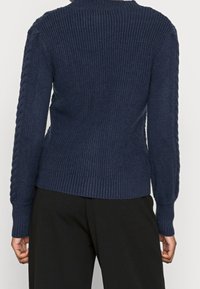 Marinblå stickad tröja med ribbad textur, med hög hals och kabelstickade detaljer på sidorna, avslutad med ribbade muddar och fåll.