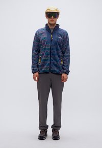 Φλις μπουφάν με μπλε και πολύχρωμα γεωμετρικά σχέδια, κλείσιμο με φερμουάρ, κίτρινες λεπτομέρειες, συνδυασμένο με γκρι παντελόνι και καφέ ορειβατικά παπούτσια.