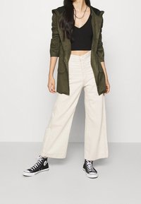 Olivgrön huva jacka, svart cropped topp, krämfärgade vida byxor och svarta sneakers. Borstad textur och synliga sömdetaljer.