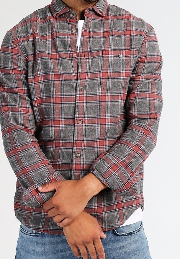 Camicia flanella a quadri rossa e grigia con colletto button-down, maniche lunghe e taschino sul petto, indossata sopra una t-shirt bianca. Vestibilità casual.
