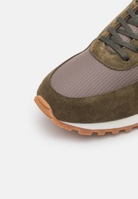 Nahaufnahme eines braunen Sneakers mit Netzobermaterial, Wildlederüberzügen, grauen Schnürsenkeln, weißer Zwischensohle und Gummi-Außensohle vor weißem Hintergrund.