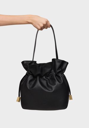 Ręka trzymająca czarną skórzaną torebkę ze ściągaczem, długimi cienkimi paskami i złotymi metalowymi zdobieniami na jednolitym tle.