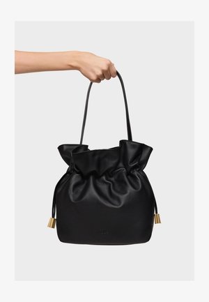 Ręka trzymająca czarną skórzaną torebkę ze ściągaczem, długimi cienkimi paskami i złotymi metalowymi zdobieniami na jednolitym tle.