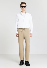Camicia bianca a maniche lunghe, t-shirt crema sotto, pantaloni beige con cordoncino, occhiali da sole neri e scarpe basse nere con lacci. Design semplice e moderno.