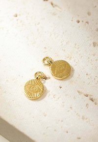 Pendente in oro dalla forma circolare, caratterizzato da design testurizzati a forma di occhio e scanalature. Pendono da piccoli anelli, poggiando su uno sfondo neutro.