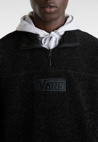 Pulóver negro de forro polar con media cremallera y superficie texturizada, con un parche del logo de Vans en el pecho y una sudadera gris llevada debajo.