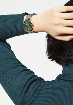 Montre ronde avec un cadran vert et des aiguilles argentées, montée sur un bracelet en maille argentée. Portée sur un poignet recouvert d'un pull en maille côtelée en teal.