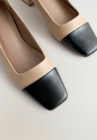 Pantofi cu vârful pătrat bej, cu capac din piele neagră, textură netedă și toc jos în formă de bloc. Design curat, modern, cu detalii minimale.