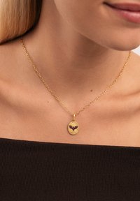 Collar de cadena de oro con un colgante ovalado que presenta un diseño de murciélago negro y pequeños acentos, reposando sobre una blusa oscura.
