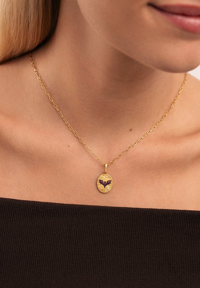 Collar de cadena de oro con un colgante ovalado que presenta un diseño de murciélago negro y pequeños acentos, reposando sobre una blusa oscura.