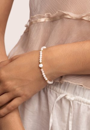 Bracciale bianco con perline, caratterizzato da perle rotonde lisce di diverse dimensioni e accenti dorati, indossato su un polso, abbinato a una blusa di tessuto leggero.