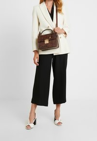 Femme portant un blazer blanc, un pantalon large noir, des talons ouverts blancs, tenant un sac à main en cuir crocodile marron avec un fermoir doré.