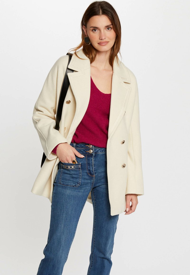 Cappotto doppiopetto in lana crema con grandi bottoni, abbinato a un top in maglia rosa intenso e jeans in denim blu con cerniere sulle tasche.