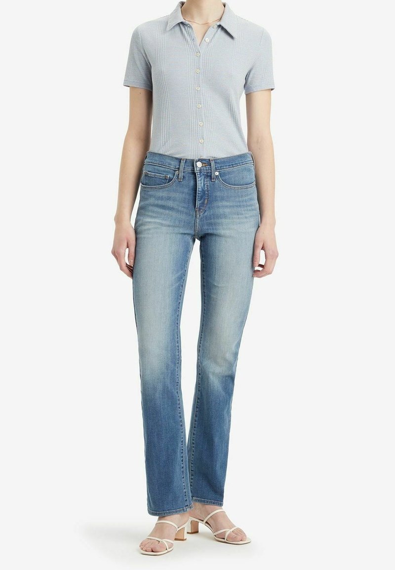 Femme portant un polo bleu clair, un jean droit délavé taille haute et des sandales à talons beiges, debout sur fond blanc.