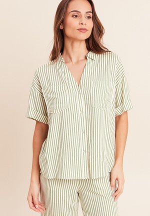 Frau trägt ein kurzärmeliges, cremefarbenes und grün gestreiftes Hemd mit Knöpfen und passende Hose, steht vor einem beigen Hintergrund.