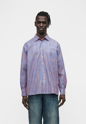 Mann trägt blau-orange gestreiftes Hemd mit Knöpfen und lockere blaue Jeans, steht vor einem schlichten hellen Hintergrund.