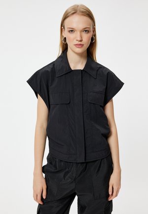 Black short-sleeve top with a structured collar, two front pockets, and a slightly textured fabric. Designed for a relaxed fit.