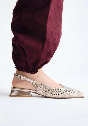 Piede che indossa una scarpa slingback beige con borchie riflettenti e tacco basso a blocchetto in metallo, abbinata a pantaloni bordeaux raccolti.