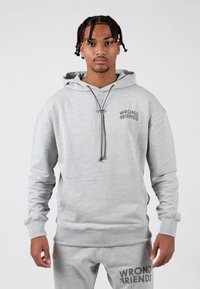 Szary hoodie z sznurkiem, w luźnym kroju, z kieszenią z przodu i napisem "WRONG FRIENDS" na klatce piersiowej. Materiał wydaje się miękki i gładki.