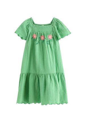 Smilende pige i en grøn kjole med blomsterbroderi og bølget kanter bærer en stråhat og brune flettede sko, midt i et skridt mod en ensartet baggrund.