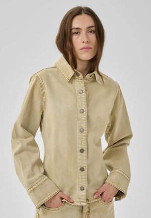 Young person with long brown hair wearing a beige button-up denim jacket and matching pants, hands in pockets, neutral background.