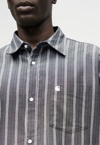 Άνδρας που φορά πουκάμισο με κάθετες ρίγες σε σκούρο γκρι και άσπρο, με κουμπιά και τσέπη στο στήθος που φέρει ένα μικρό λευκό λογότυπο.