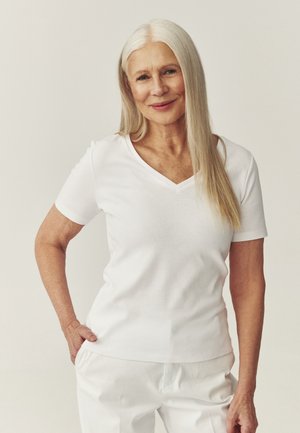 Ældre kvinde med langt hvidt hår smiler, iført en hvid V-hals trøje og hvide bukser, stående foran en ensfarvet beige baggrund.
