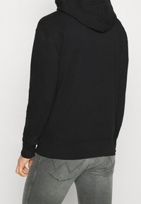 Sudadera negra con un gran bolsillo trasero, puños y dobladillo acanalados. Tejido suave con una textura lisa. Llevada sobre unos jeans grises con detalles en patrón.