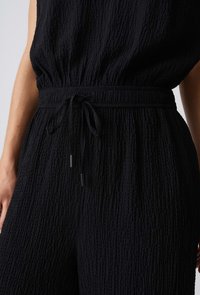 Combinaison en tissu noir texturé avec taille élastique et cordon noué, montrée du milieu du torse jusqu'aux cuisses supérieures sur une personne.