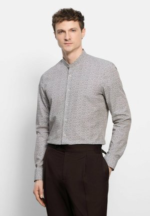 Mann trägt ein langärmliges, gemustertes Hemd mit Mandarinkragen und dunkle Hose, steht vor einem einfarbigen hellen Hintergrund.