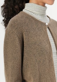 Brown cardigan made of soft fabric, featuring a round neck, a single button closure, and worn over a striped turtleneck top.