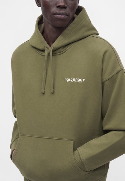 Άνδρας που φοράει φούτερ Polo Sport Ralph Lauren σε ελιά πράσινη απόχρωση με μπροστινό τσεπάκι και κορδόνια, τα χέρια εν μέρει στις τσέπες, σε απλό φόντο.