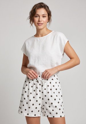 Vrouw draagt een wit kortärmelig topje en witte shorts met zwarte stippen, ze stelt het trekkoord van de shorts af.
