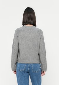 Grå strikkegenser med ribbestrikket hem og mansjetter, med raglanermer. Myk tekstur og avslappet passform, brukt sammen med blå jeans.
