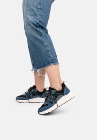 Blaue und schwarze sportliche Sneaker mit strukturiertem Stoff, kontrastierenden Akzenten und weißer Sohle. Die Schnürsenkel sind schwarz mit reflektierenden Details.