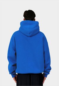 Felpa blu con un grande cappuccio, vestibilità comoda e polsini a costine. Il materiale sembra morbido con una finitura testurizzata. Pantaloni neri plissettati visibili in basso.
