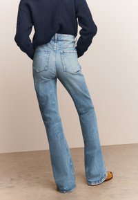 Platėjančios šviesiai mėlynos spalvos džinsinės kelnės su aukštu liemeniu, turi du galinius kišenes ir išblukusią tekstūrą. Dėvimos su tamsiai megztu megztiniu.